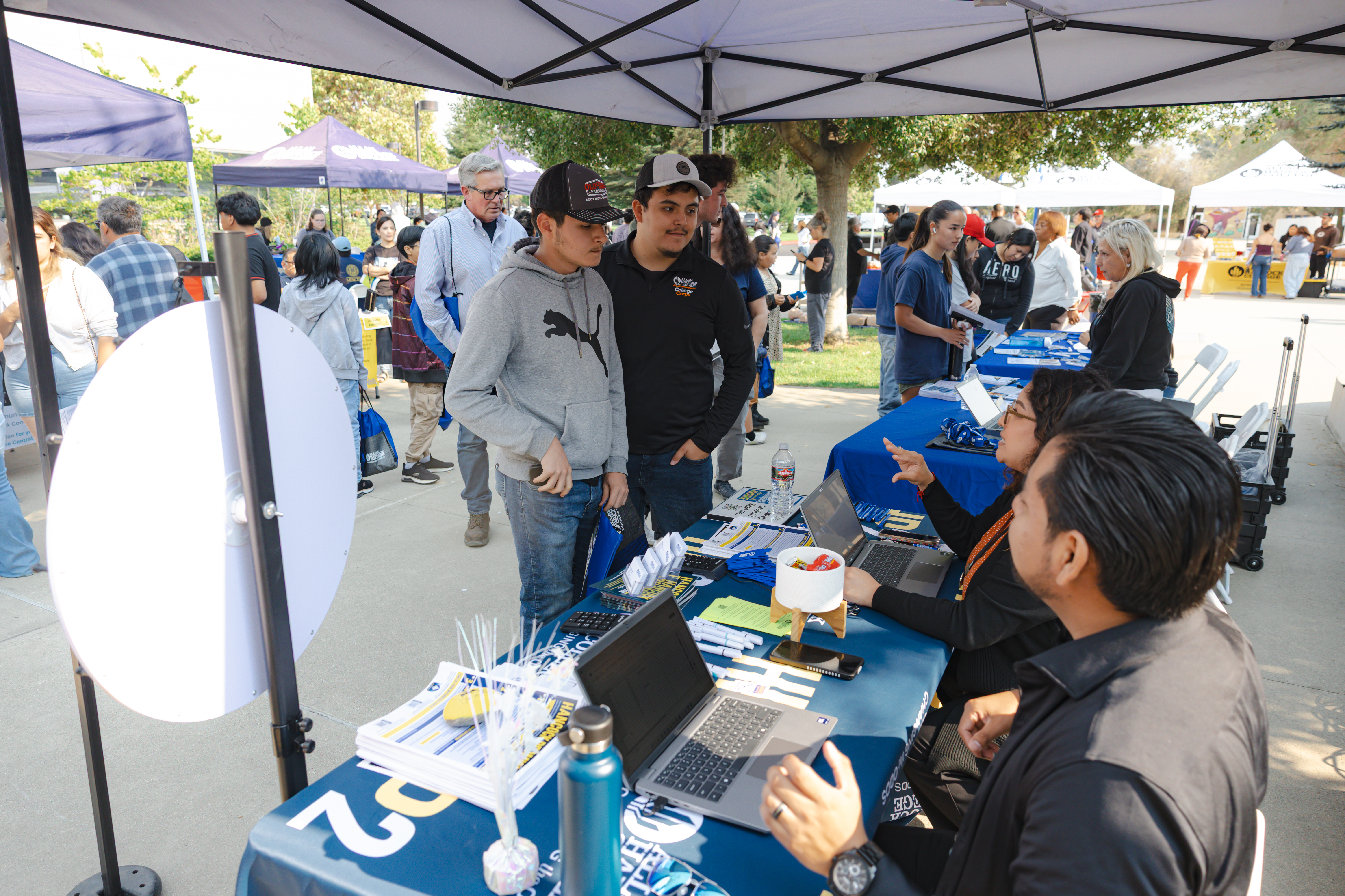 Students speak with Allan Hancock College staff at an information table during a Hancock Hello orientation event. The outdoor event features campus resource booths, laptops, brochures, and giveaway items, with students and staff gathered under canopies across the busy campus walkway.